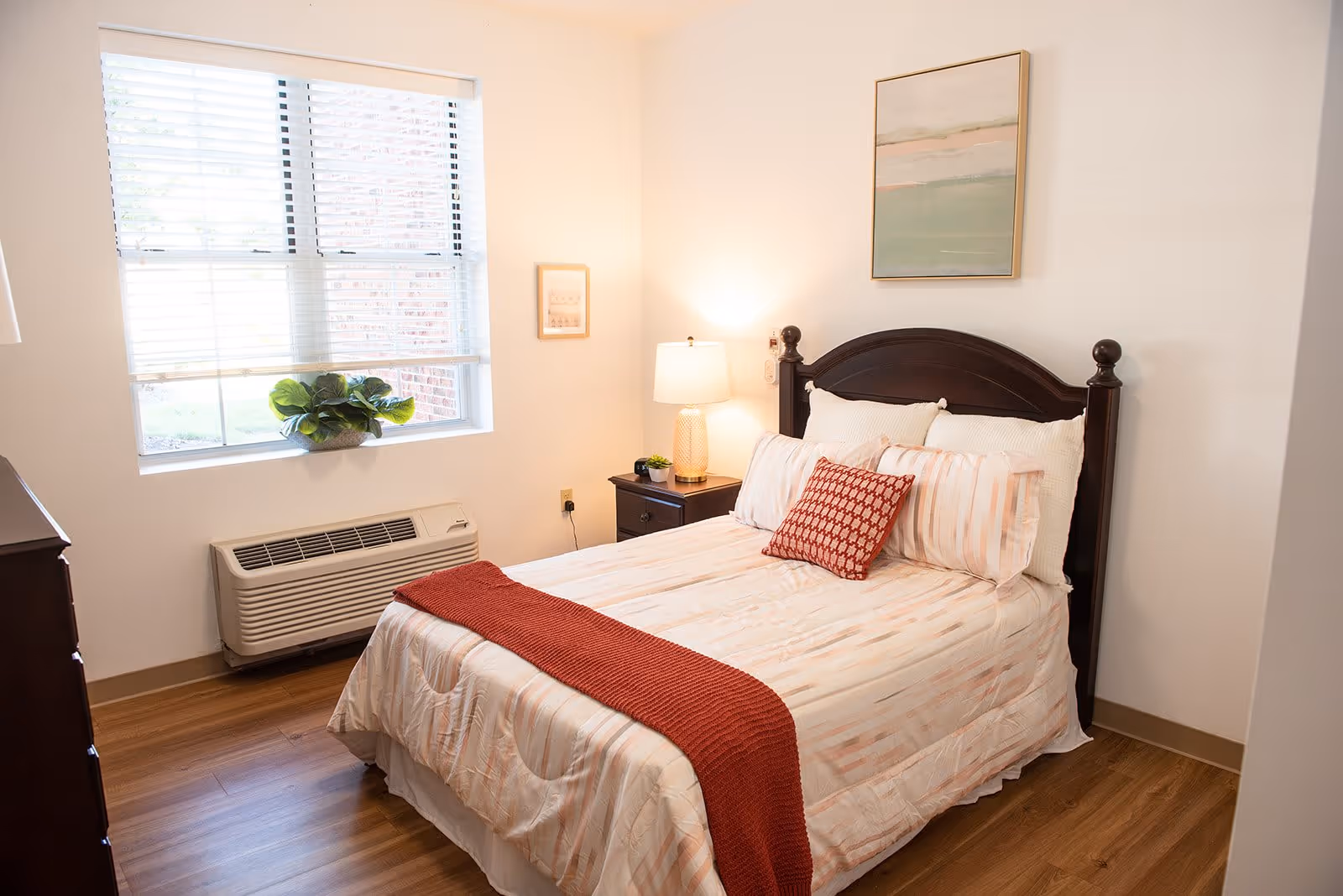 A cozy bedroom with a wooden bed frame, a neatly made bed with light-colored bedding and a red throw blanket and pillow. There is a nightstand with a lamp and a small plant, a window with blinds letting in natural light, and a framed artwork on the wall above the bed.