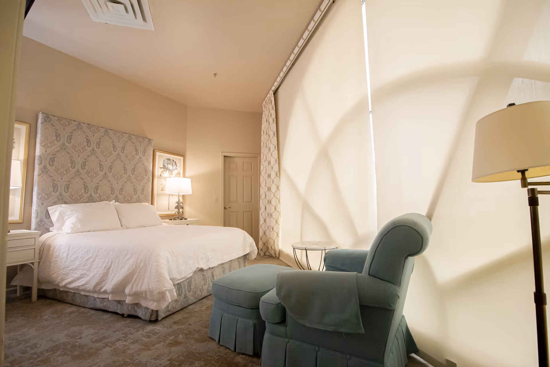A cozy bedroom with a large bed featuring a patterned headboard and white bedding. There are two bedside tables with lamps and framed artwork on the walls. A comfortable blue armchair with an ottoman is positioned near a window covered with light-colored curtains. A floor lamp stands next to the armchair.