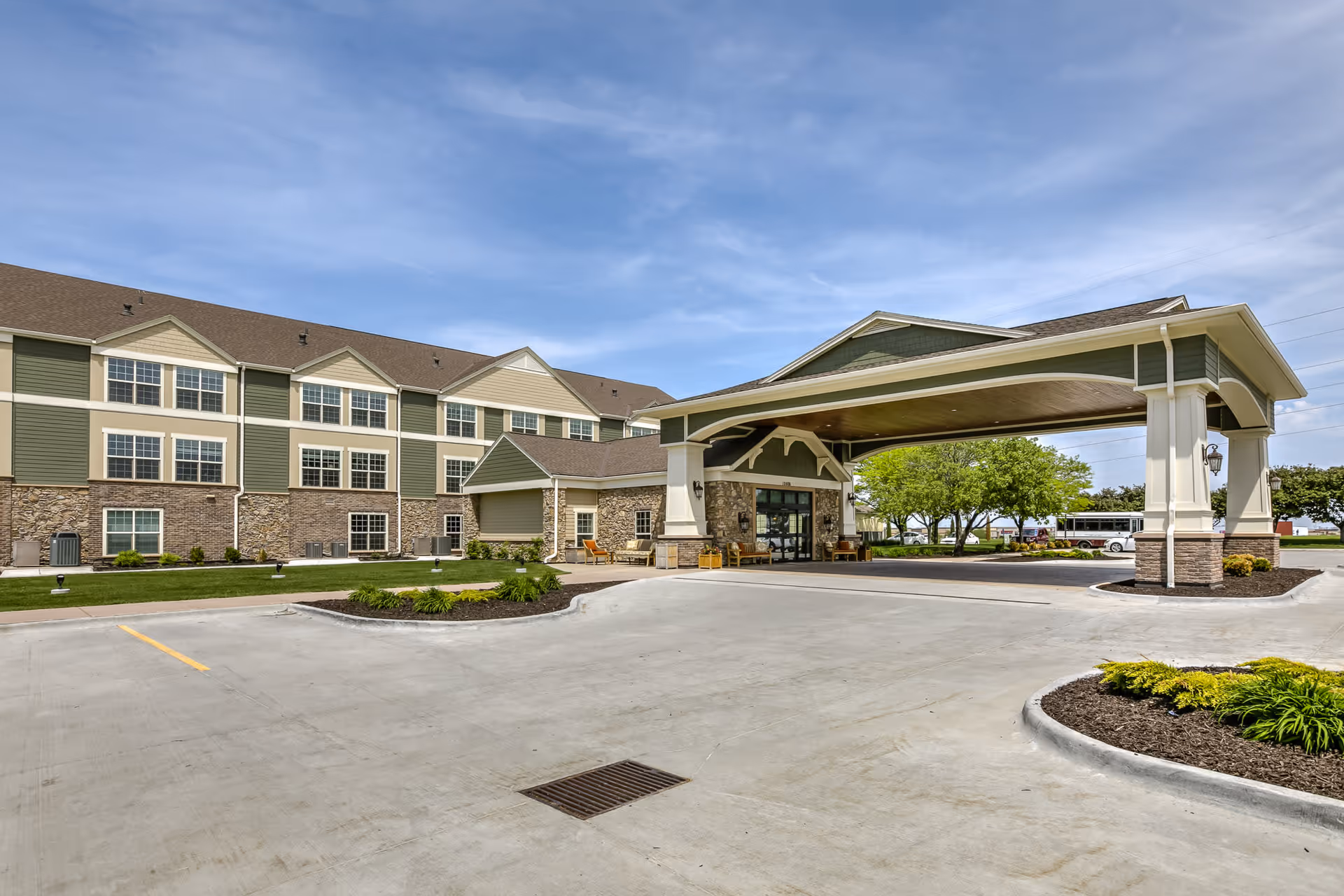 Exterior view of Remington Heights Retirement Community showing a large covered entrance with seating, a three-story building with green and beige siding and stone accents, a parking area, and landscaped greenery under a partly cloudy sky.