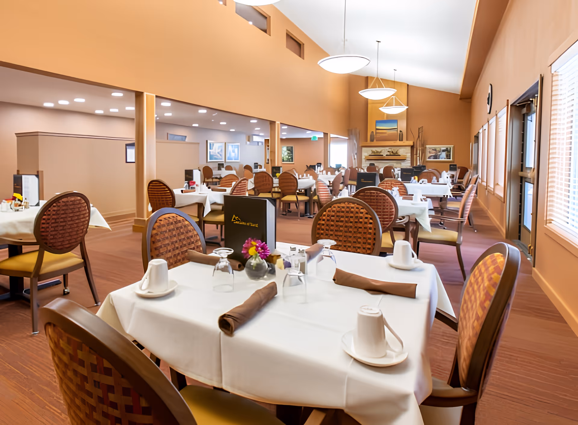 A spacious dining room in a retirement community with multiple tables covered in white tablecloths, each set with cups, glasses, napkins, and small flower arrangements. The room features warm-toned walls, large windows with blinds, and modern hanging light fixtures. Comfortable wooden chairs with patterned backs surround the tables, and a fireplace is visible in the background.