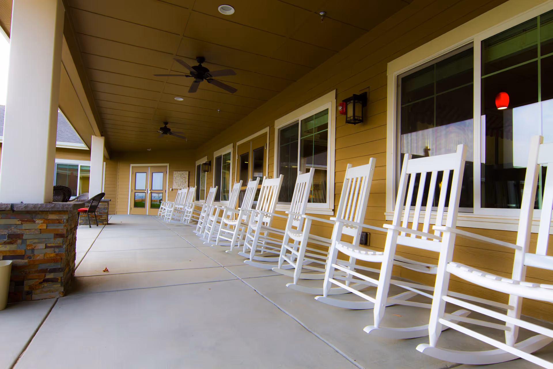 A covered outdoor patio area with a row of white rocking chairs lined up against the wall of a building with large windows. The patio has ceiling fans and stone pillars, creating a comfortable seating space.