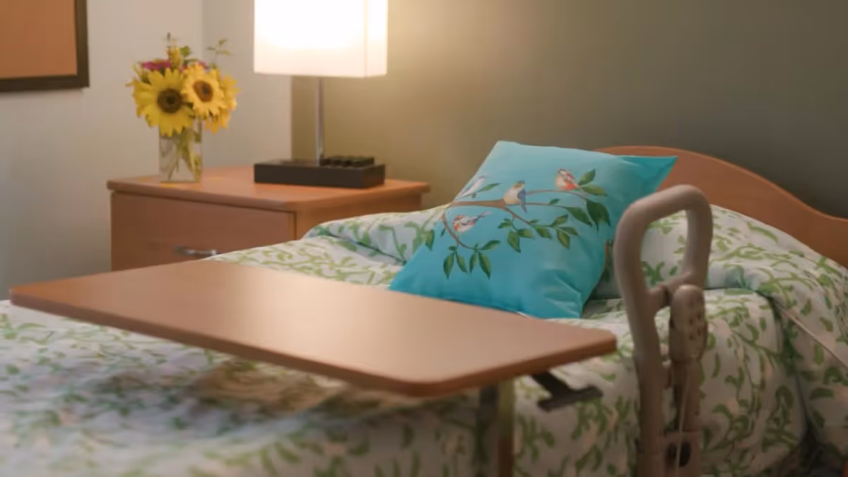 A neatly made bed with green and white patterned bedding and a blue pillow featuring a bird design. There is a wooden overbed table positioned over the bed. Next to the bed is a wooden nightstand with a lamp and a vase of sunflowers. The wall behind the bed is painted a muted green color.