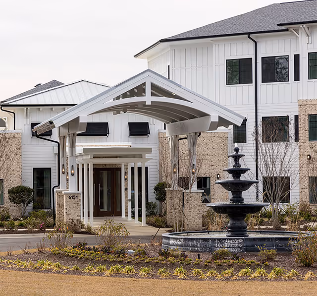 Front entrance of a white senior living building with a covered porte-cochere and a multi-tiered fountain in the landscaped forecourt.