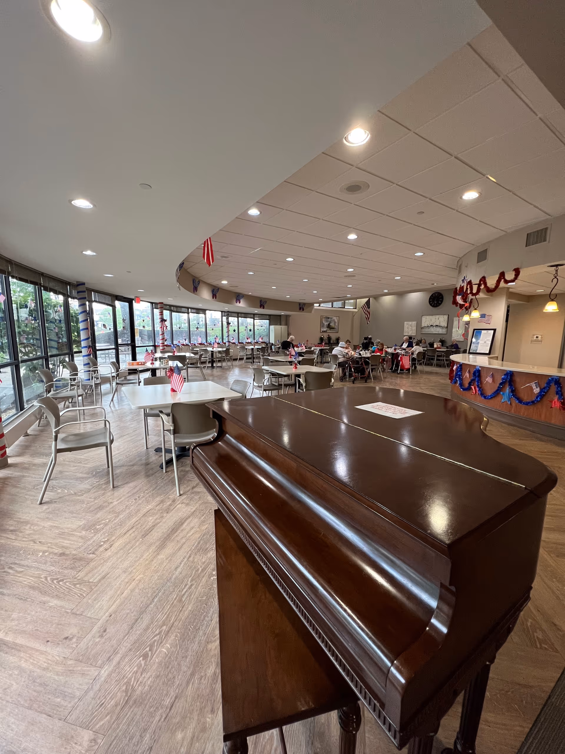 A senior facility dining room with a grand piano in the foreground, rows of tables and chairs, and patriotic decorations.