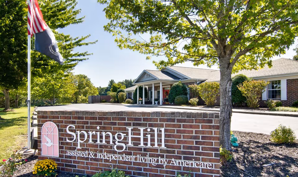 Exterior view of a senior living facility with a brick sign in the foreground that reads 'SpringHill assisted & independent living by Americare'. The building is a single-story structure with a covered entrance, surrounded by trees and landscaping under a clear blue sky.