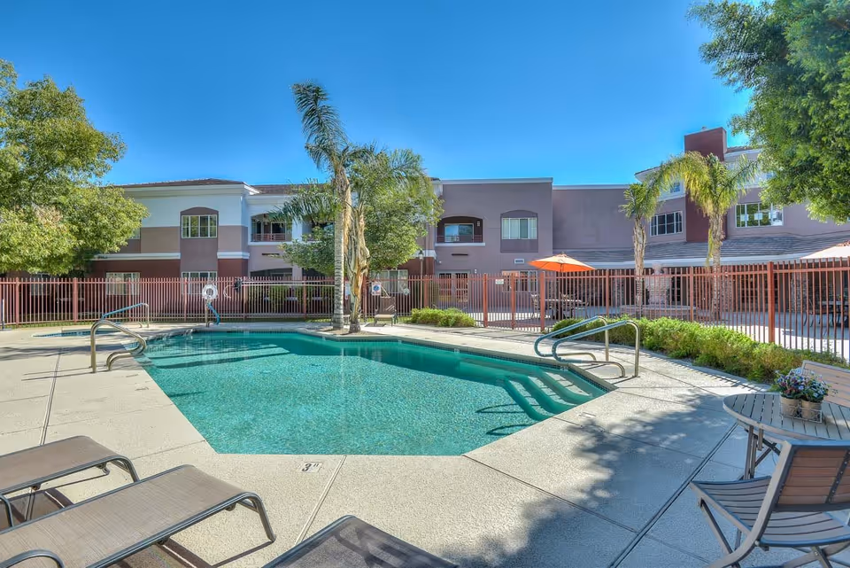 Outdoor swimming pool area at Chaparral Winds Assisted Living with lounge chairs, palm trees, and a fenced courtyard surrounded by a two-story building under a clear blue sky.