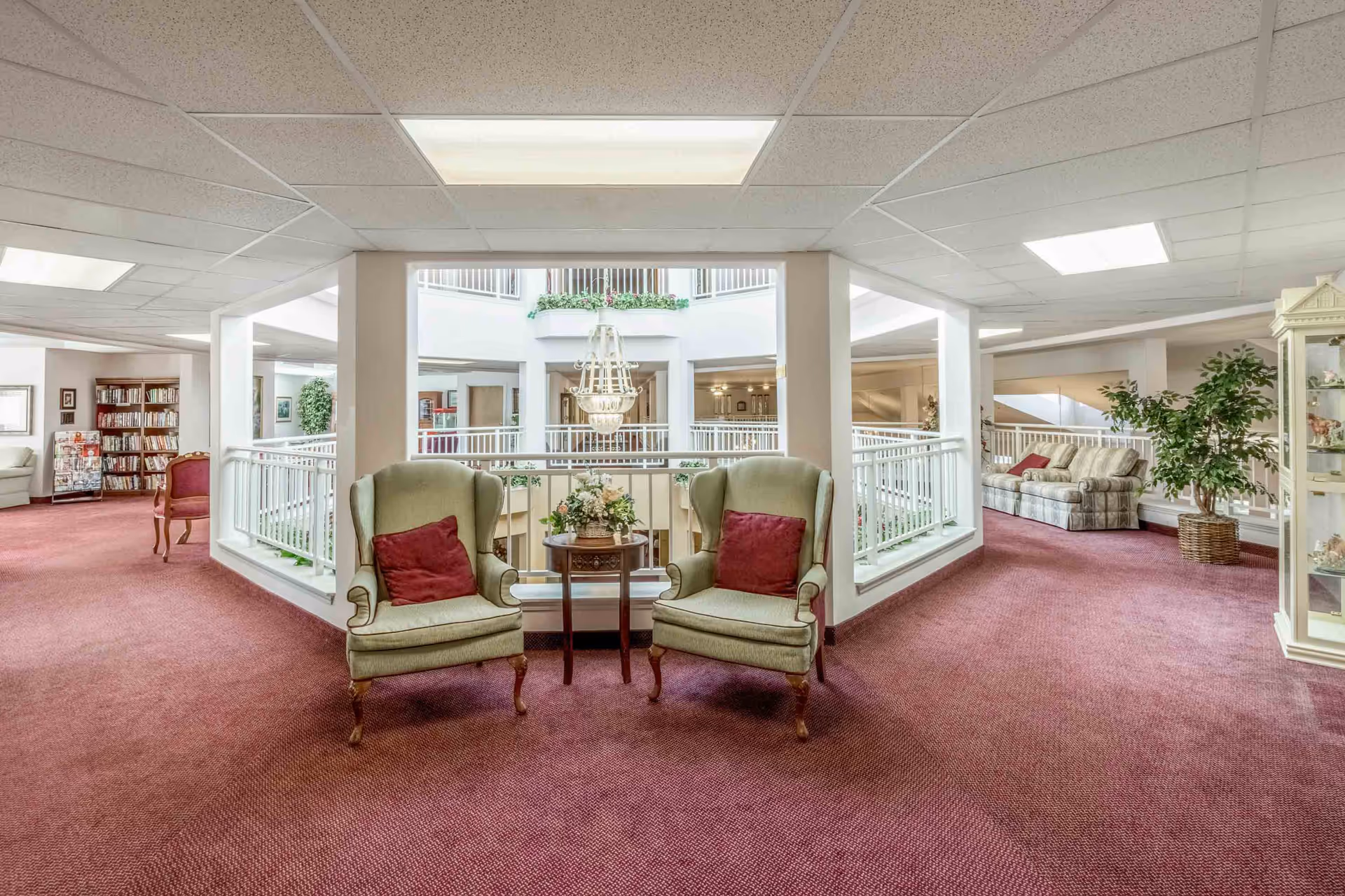 Carpeted common lounge with two upholstered armchairs and a small table overlooking an atrium with a chandelier and additional seating and bookshelves.