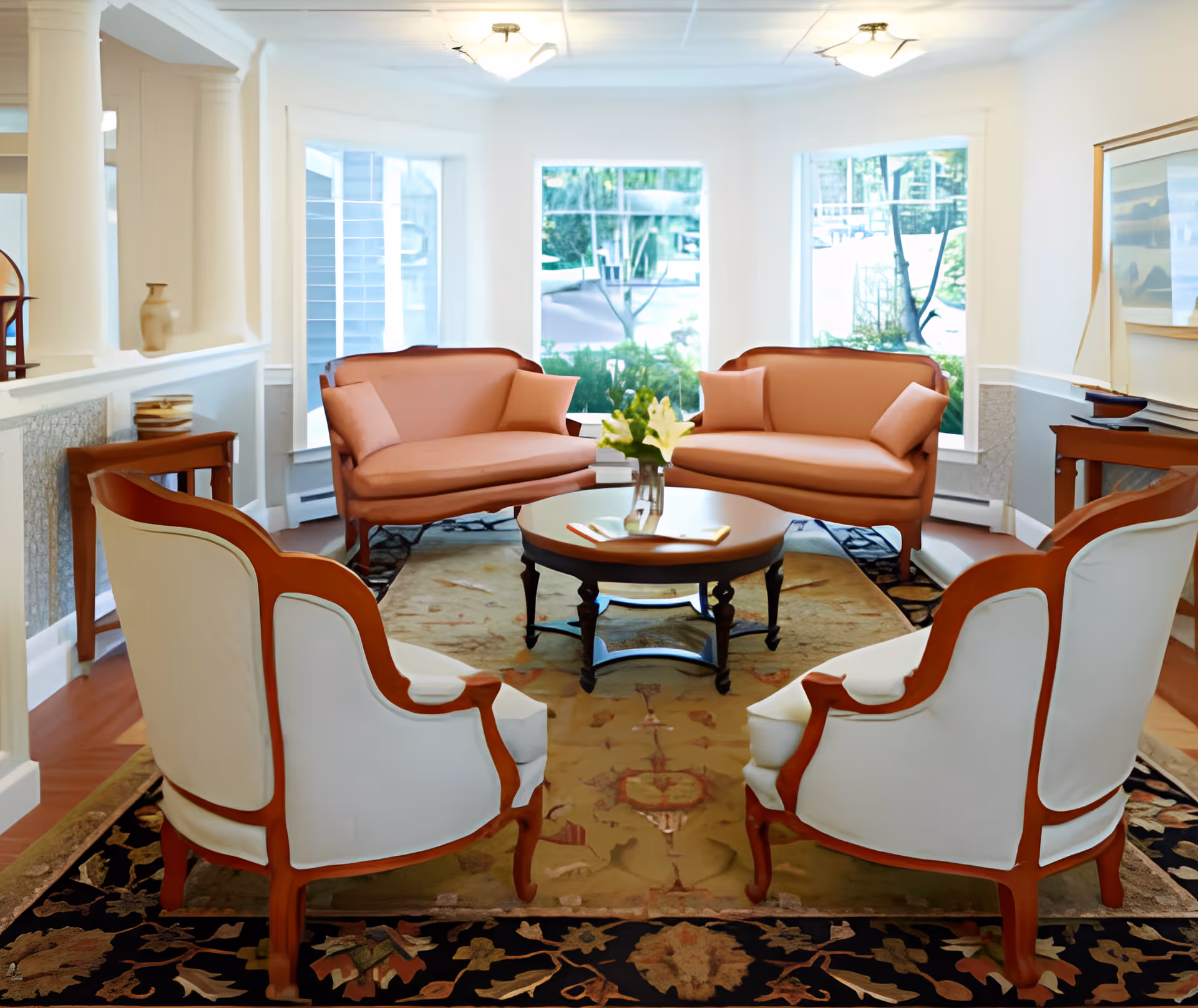 A bright and cozy living room with two pink upholstered loveseats, two white armchairs with wooden frames, a round wooden coffee table with a vase of flowers, and large windows letting in natural light. The room features a patterned area rug and light-colored walls with decorative molding.