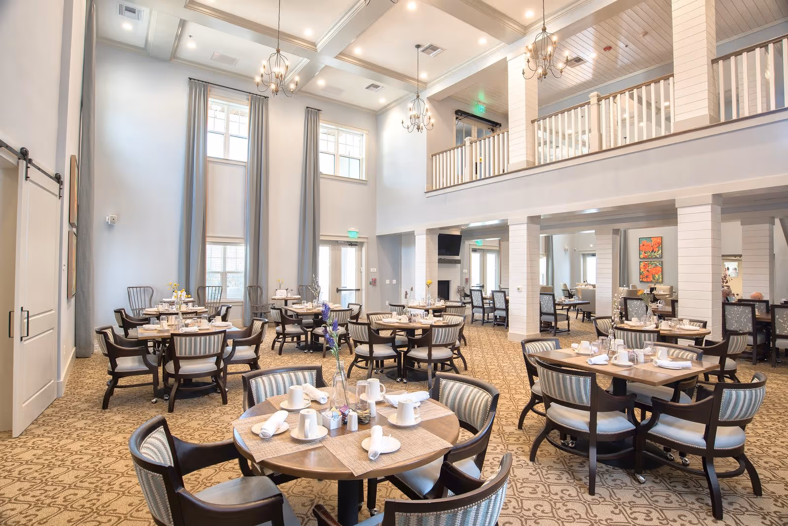 A spacious and elegant dining room with multiple round wooden tables set with white cups, napkins, and placemats. The room features high ceilings with chandeliers, tall windows with long curtains, and a second-floor balcony overlooking the area. The carpet has a patterned design, and the overall decor is bright and inviting.
