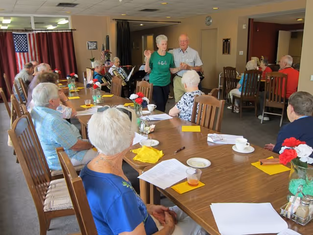 A group of elderly people seated around a large wooden table in a communal room, with some individuals dancing and others playing an accordion. The room has an American flag on the wall and is decorated with flowers on the table.