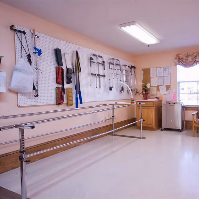A rehabilitation room with parallel bars for walking exercises, a pegboard on the wall holding various mobility aids such as walkers, braces, and slings. The room has light peach walls, a window with a floral valance, a small cabinet with a plant and papers pinned on a bulletin board, and a small refrigerator.