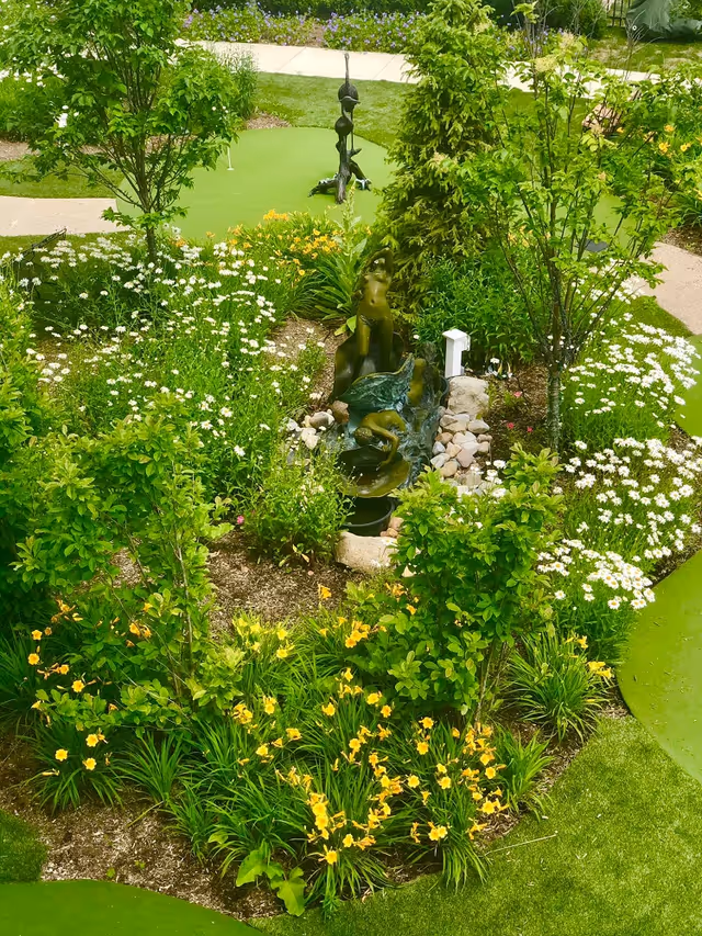 A lush garden area featuring various green shrubs, yellow and white flowers, and a small bronze statue of a woman surrounded by rocks. The garden is bordered by a green putting green with a golf hole and flag visible in the background.