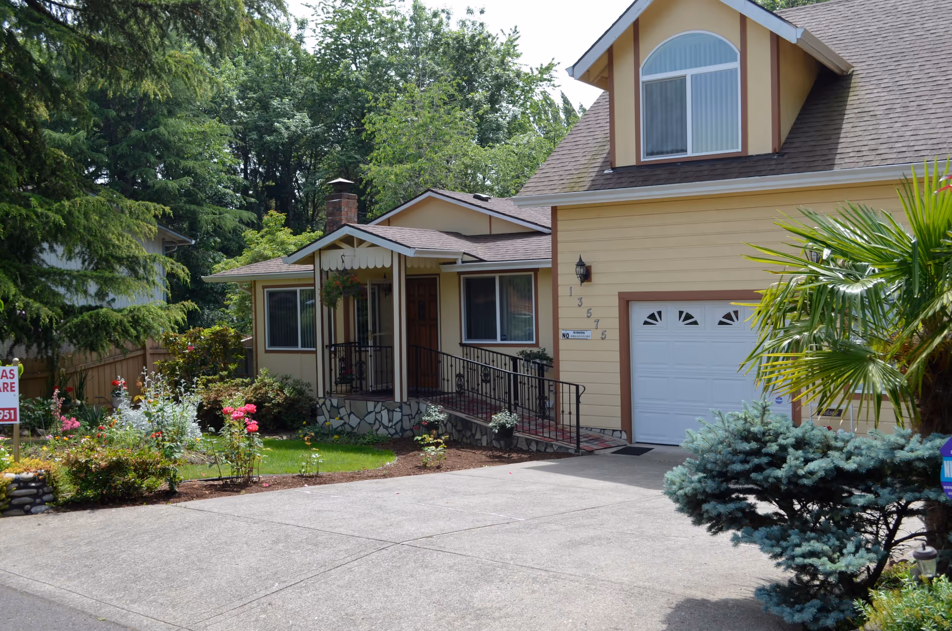Exterior view of a yellow two-story house with a garage, a small porch with a ramp, and a well-maintained garden with various plants and flowers. The house is surrounded by trees and greenery.