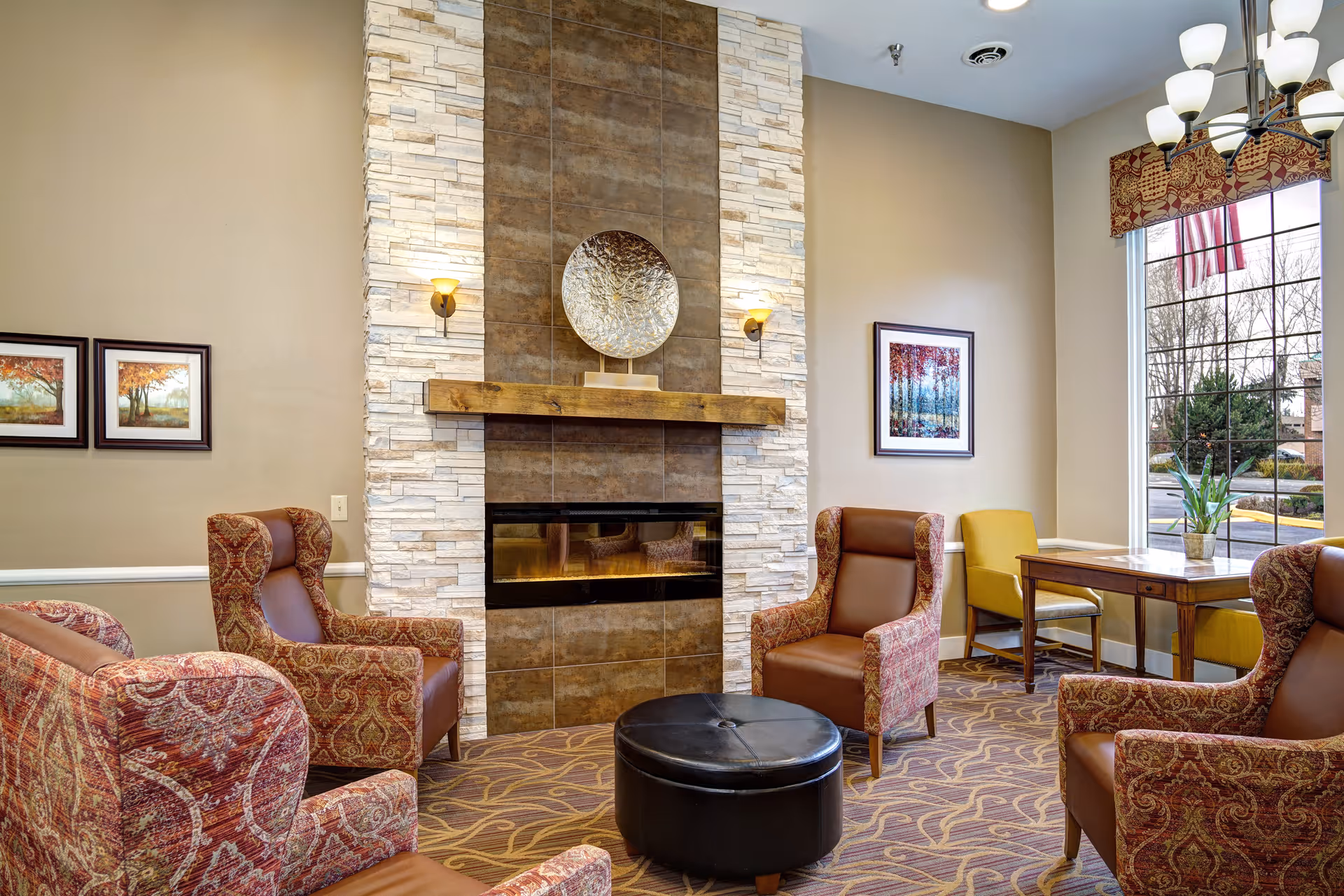 A cozy living room area with four patterned armchairs arranged around a black round ottoman in front of a modern stone fireplace. The room has beige walls, framed artwork, a large window with a valance, and a wooden table with two yellow chairs near the window.