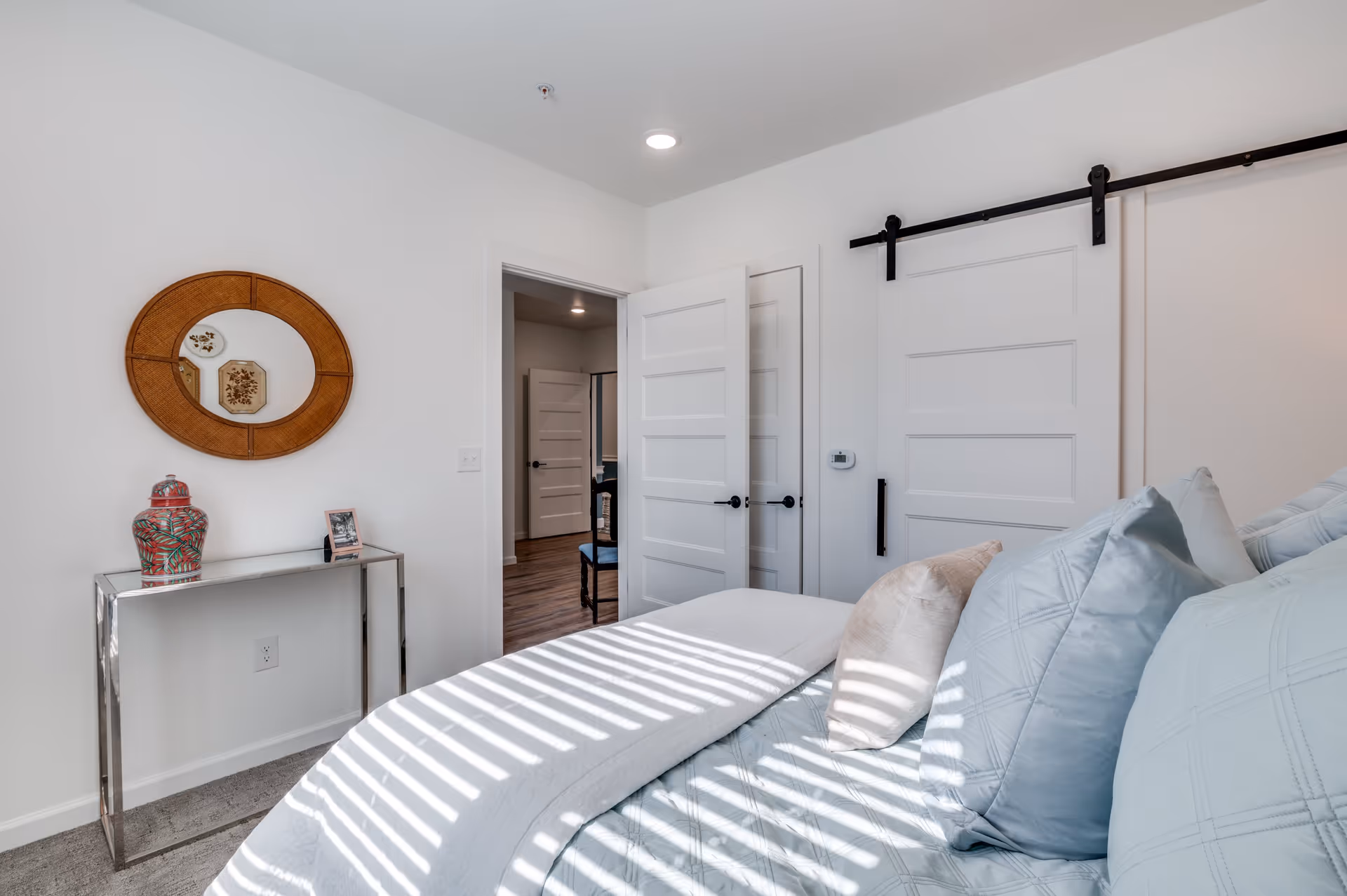 Sunlit bedroom with a neatly made bed, decorative pillows, a console table with a vase, a round wall mirror, and white sliding barn doors.
