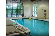 Indoor swimming pool in a bright room with large arched windows, pool toys on the edge and lounge chairs nearby.
