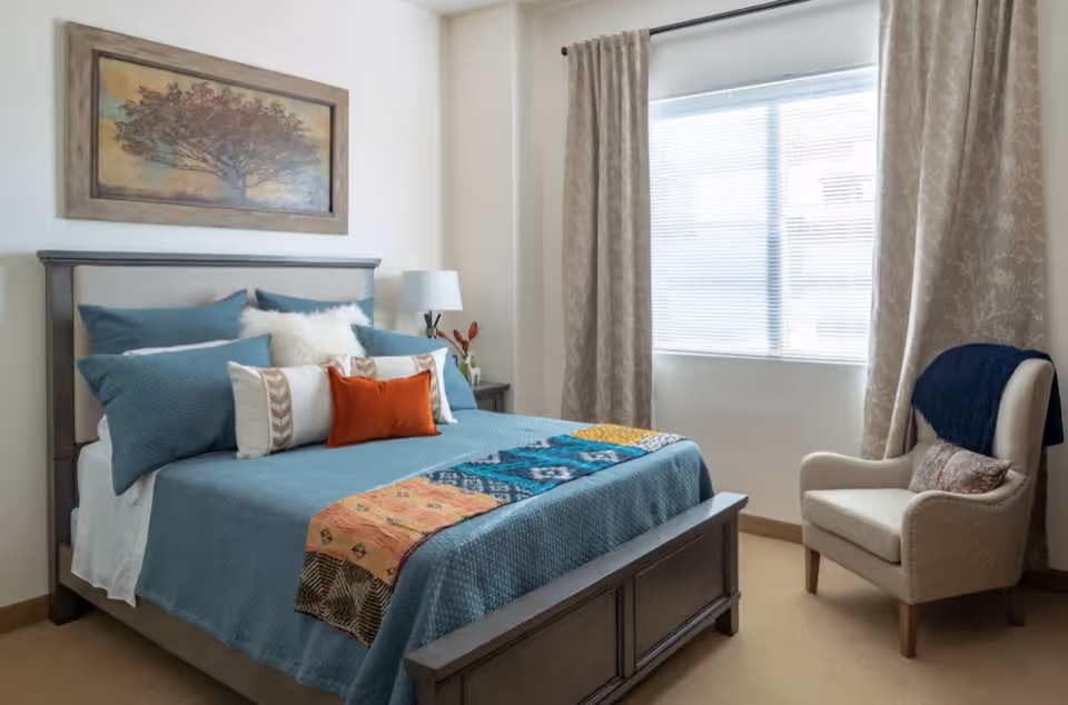 A neatly made bed with blue bedding and multiple pillows in a bedroom. There is a colorful quilt at the foot of the bed. A framed picture of a tree hangs above the bed. To the right, there is a window with beige patterned curtains and a beige armchair with a dark blue throw draped over the back.