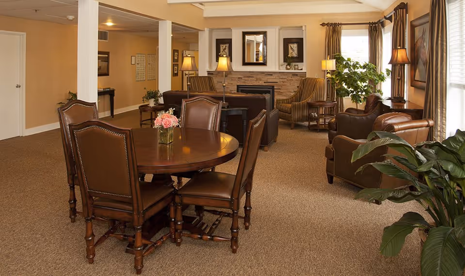 A cozy senior living common area featuring a round wooden table with six leather chairs, a fireplace with a stone surround, two armchairs, a sofa, several lamps, framed artwork on the walls, and large windows with curtains letting in natural light. There are also green plants adding a touch of nature to the warm, inviting space.