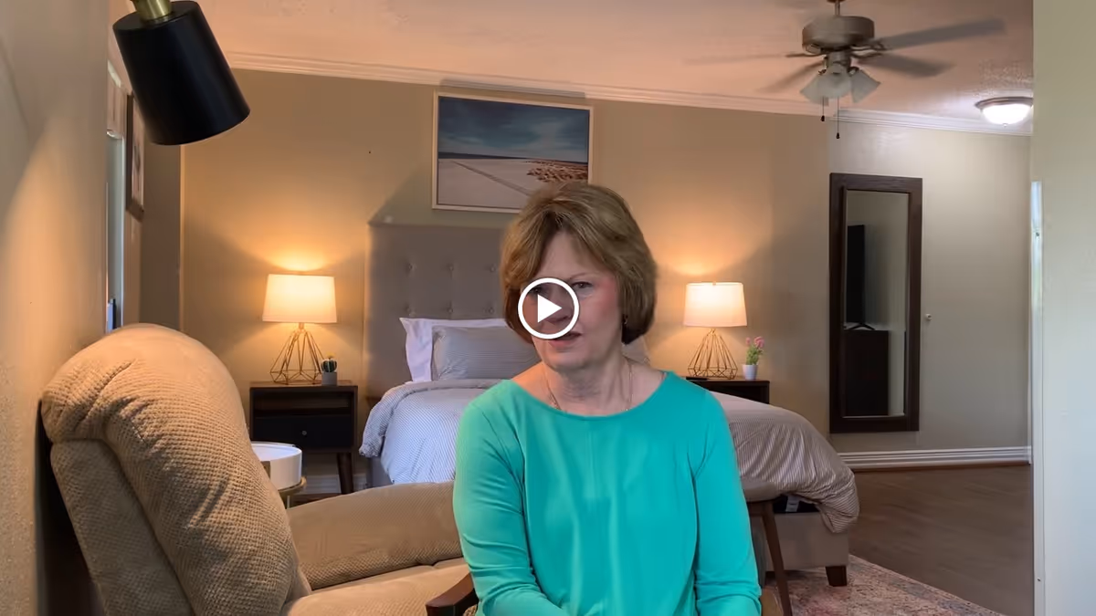 A woman in a green top sitting in a beige armchair in a bedroom with a bed, two bedside tables with lamps, a ceiling fan, a wall mirror, and a framed picture above the bed.