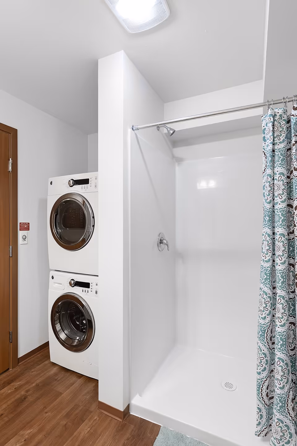 A bathroom area featuring a white shower stall with a floral patterned shower curtain on the right and a stacked washer and dryer unit on the left. The floor is wood, and the walls are white with a wooden door partially visible.