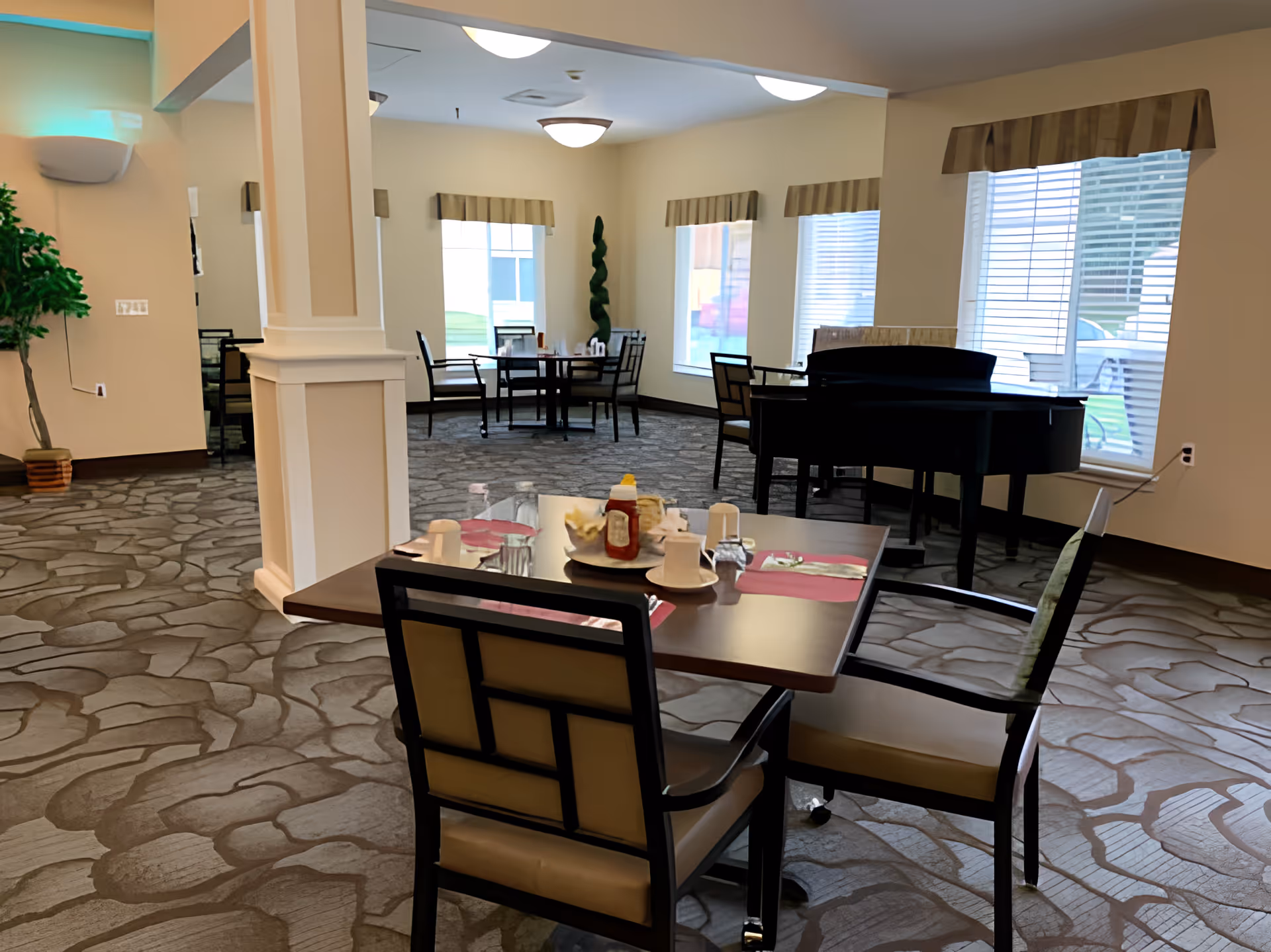 Bright communal dining room with set tables and chairs, a piano by the windows, and decorative columns.
