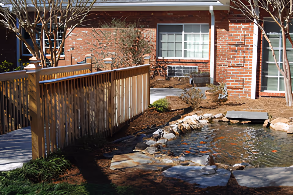 Landscaped courtyard with a wooden footbridge over a koi pond in front of a brick assisted-living building.