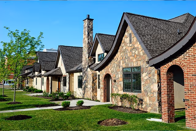 Row of charming single-story residential buildings with stone and brick facades, steeply pitched roofs, and well-maintained green lawns and small trees under a clear blue sky.