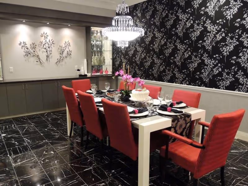 A dining room with a long white table set for eight people with red upholstered chairs. The table is decorated with a floral centerpiece, a cake, and place settings with black napkins. The room has black marble flooring, a black and white floral wallpaper on one wall, and a chandelier hanging above the table. There is a cabinet with decorative items and a wall decoration with metal leaves on the opposite wall.
