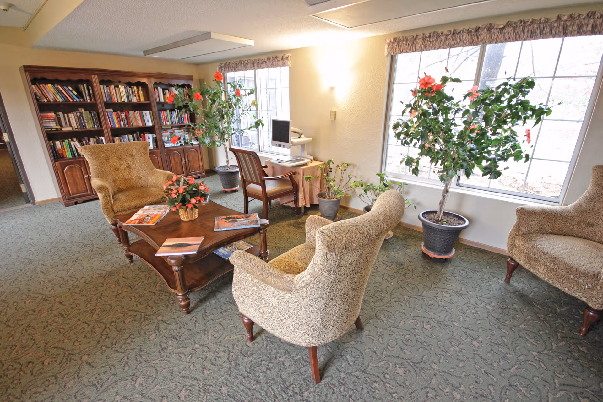 Cozy sitting area with upholstered armchairs around a wooden coffee table, bookshelves, potted plants, and a desk by large windows.