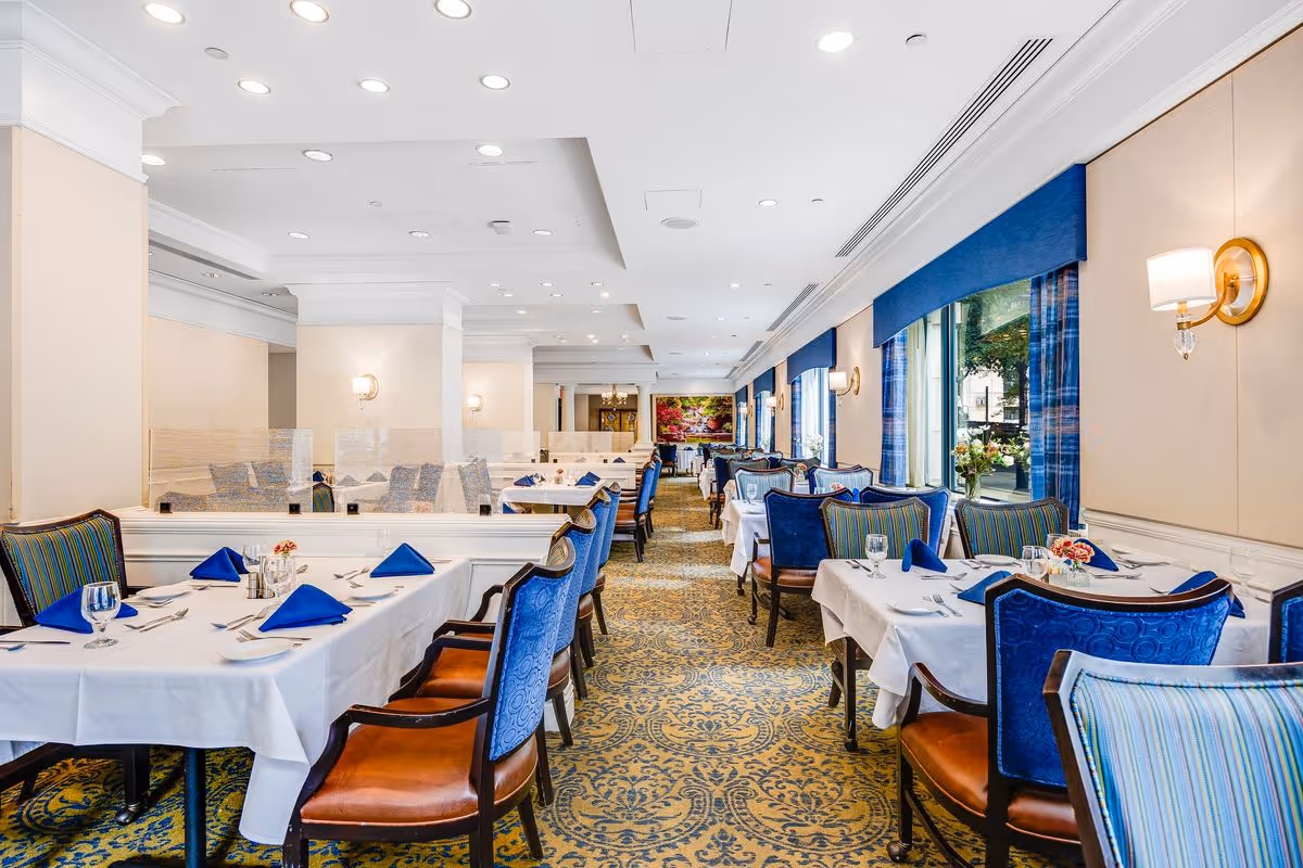 A bright and elegant dining room with tables covered in white tablecloths, set with blue napkins, glassware, and silverware. The room features blue upholstered chairs, patterned carpet, large windows with blue curtains, and wall sconces providing warm lighting.