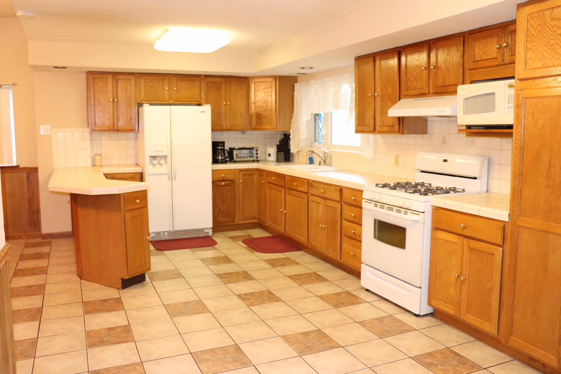 A spacious kitchen with wooden cabinets, a white refrigerator, a white gas stove with oven, a white microwave, and a double sink under a window with lace curtains. The floor is tiled with a pattern of beige and brown squares, and there are two red floor mats in front of the refrigerator and sink. The kitchen has a tiled countertop and a small peninsula with additional cabinet storage.