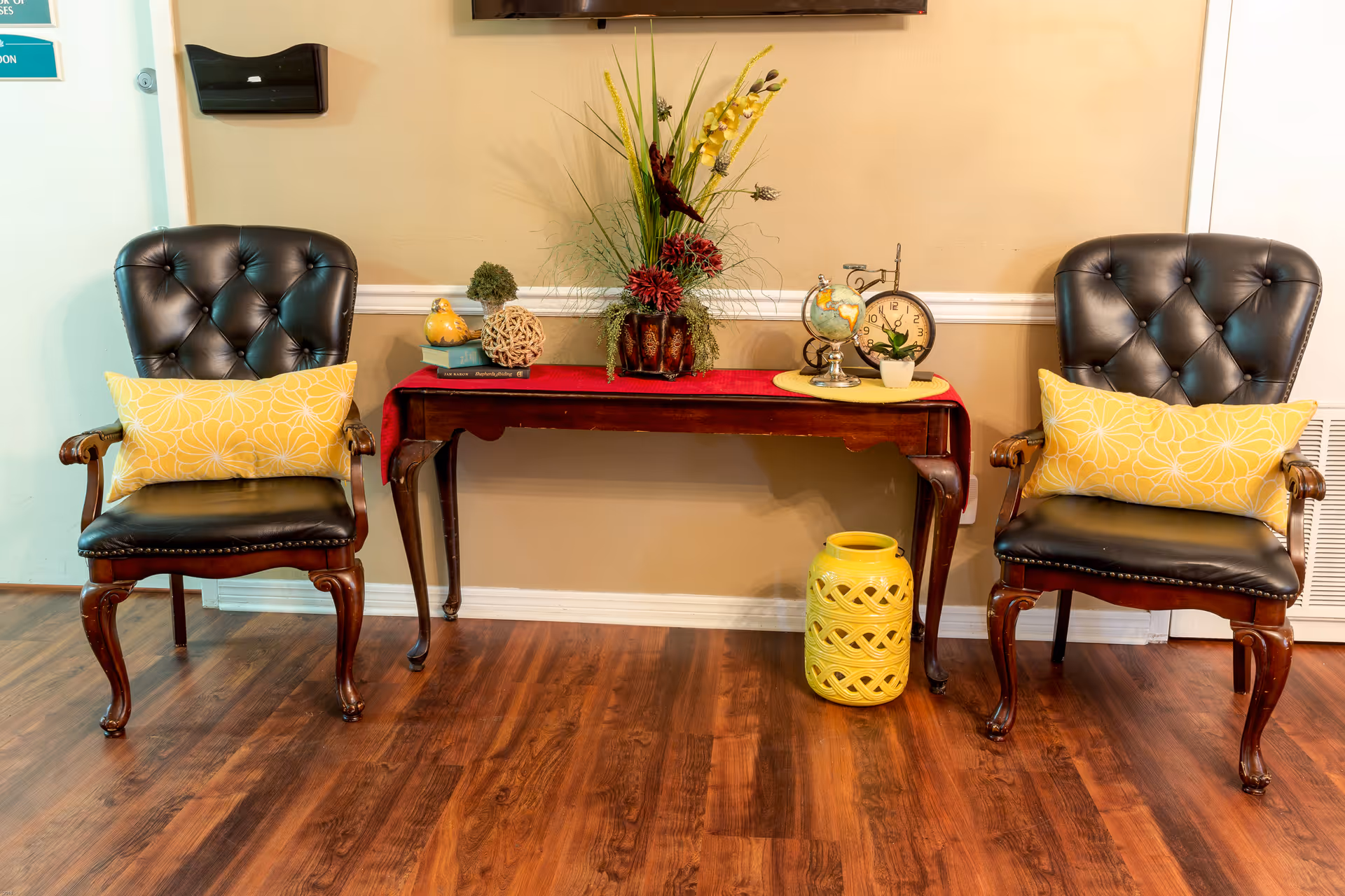 Two black leather tufted armchairs with wooden legs and yellow patterned cushions flank a wooden console table with a red runner. On the table are decorative items including a globe, clock, small plant, and a floral arrangement. A yellow decorative ceramic stool is placed on the floor beneath the table. The room has wood flooring and beige walls.
