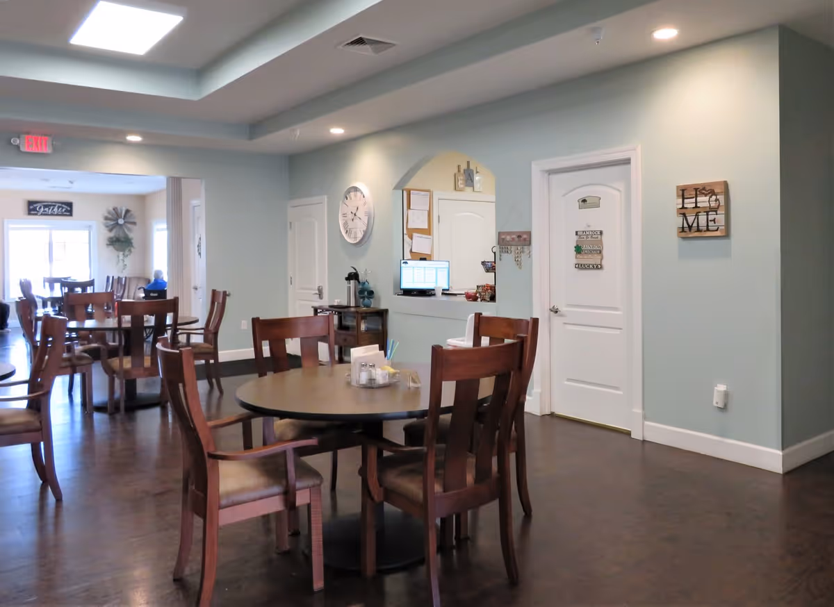 A dining area in a senior living facility with round wooden tables and chairs. The room has light blue walls, a large wall clock, and decorative signs on the walls. There is a small counter with a computer and coffee station, and an exit sign is visible in the background near another room with more tables and chairs.
