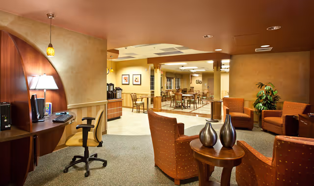 Interior view of a senior living facility common area with a computer workstation on the left, several wooden tables and chairs in the background, and a seating area with four orange upholstered armchairs around a small wooden table with decorative vases. The walls are painted in warm tones and there is a potted plant in the corner.