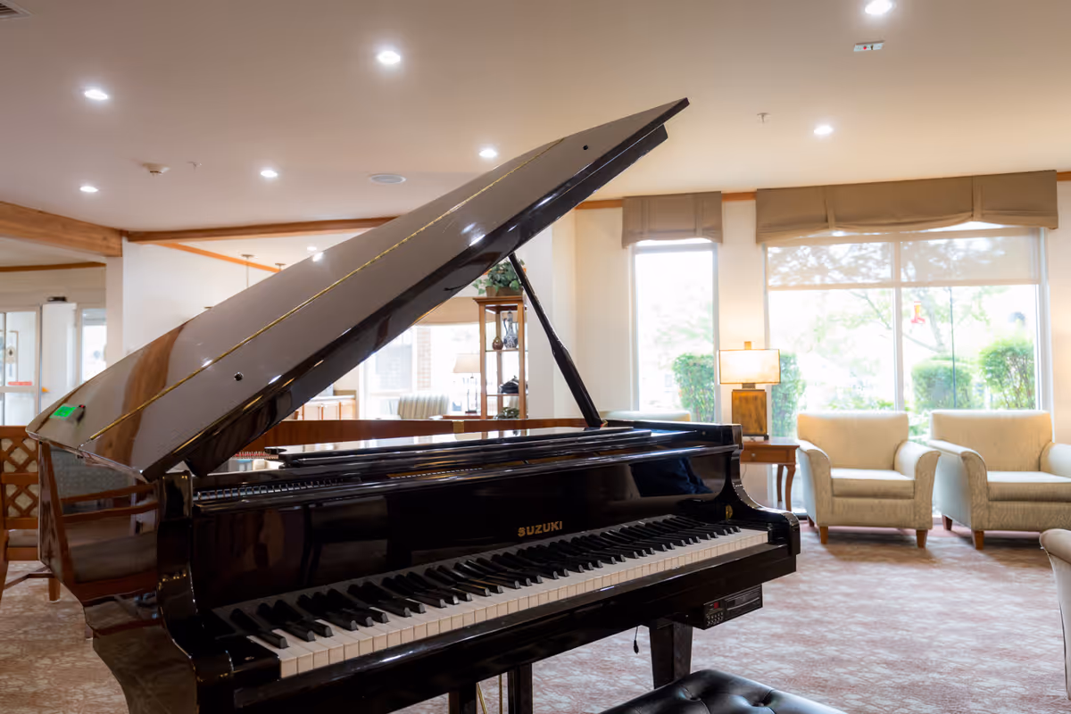 A black grand piano with the lid open in a well-lit living room area featuring beige armchairs, a side table with a lamp, large windows with beige valances, and a carpeted floor.