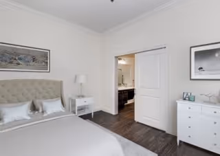 Bright bedroom with a bed, nightstand, dresser, and an open double door revealing a bathroom vanity area.