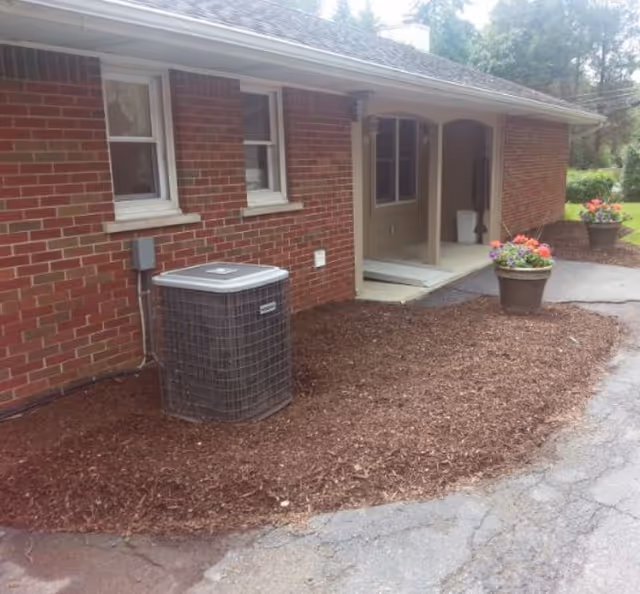 Exterior side view of a brick building with two windows, an air conditioning unit, and a covered entrance. There are two large flower pots with colorful flowers placed on a mulched area next to a paved driveway.