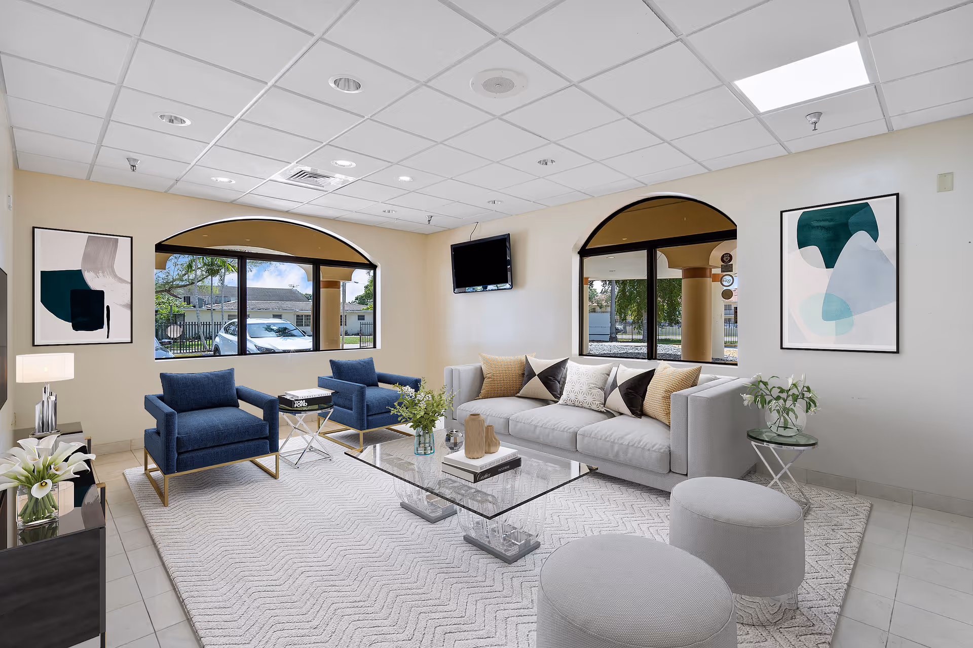 A bright and modern living room with a light gray sofa adorned with patterned pillows, two blue armchairs with gold legs, a glass coffee table on a light patterned rug, two round gray ottomans, and two abstract art paintings on the walls. Large arched windows provide natural light and views of the outside. A flat-screen TV is mounted on the wall.