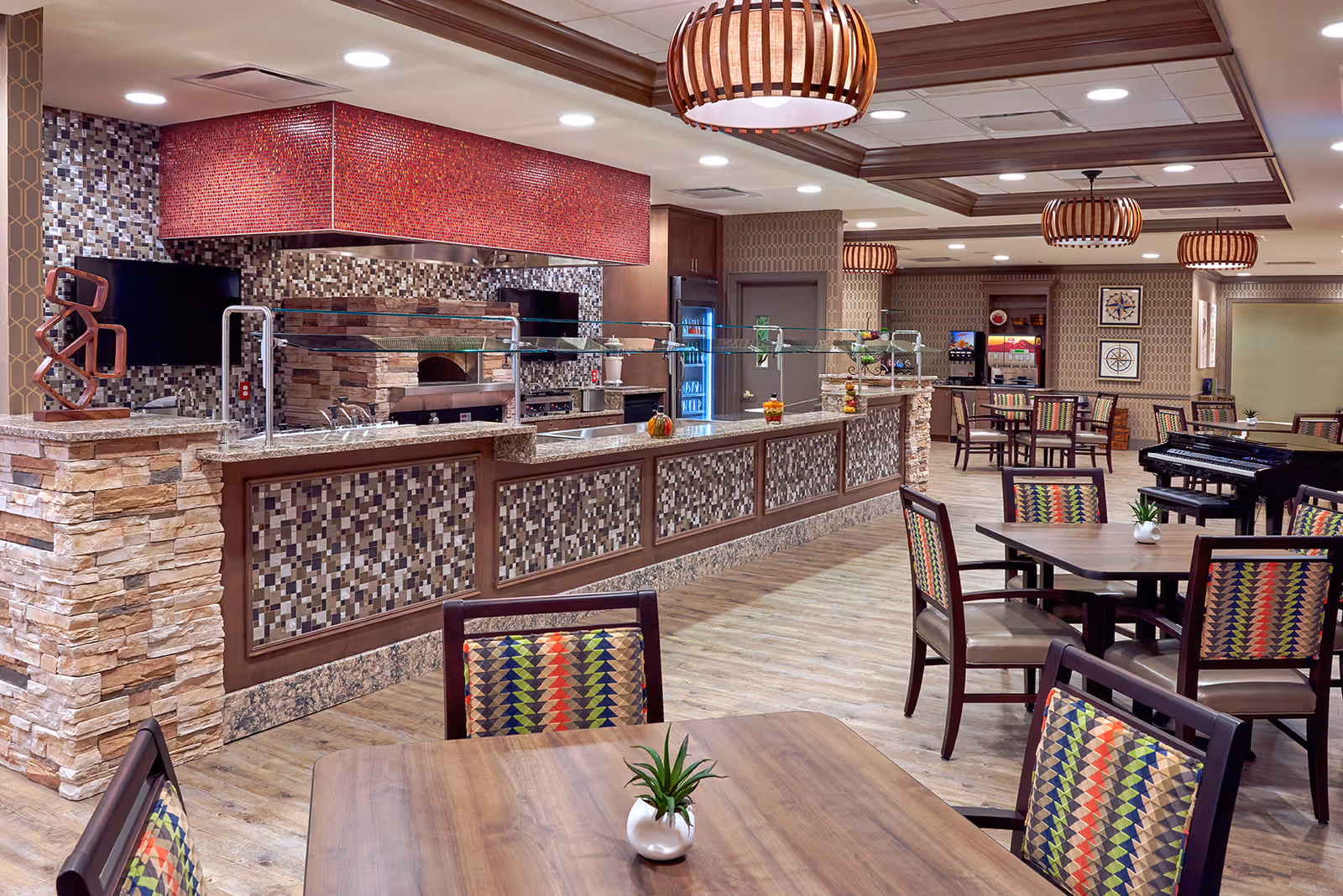 A spacious dining area in an assisted living facility featuring a long serving counter with a stone and mosaic tile facade, overhead red mosaic hood, and glass sneeze guards. The room has multiple wooden tables and chairs with colorful patterned upholstery, wooden ceiling beams, and modern pendant lights. A piano is visible in the background along with beverage dispensers and framed artwork on the walls.