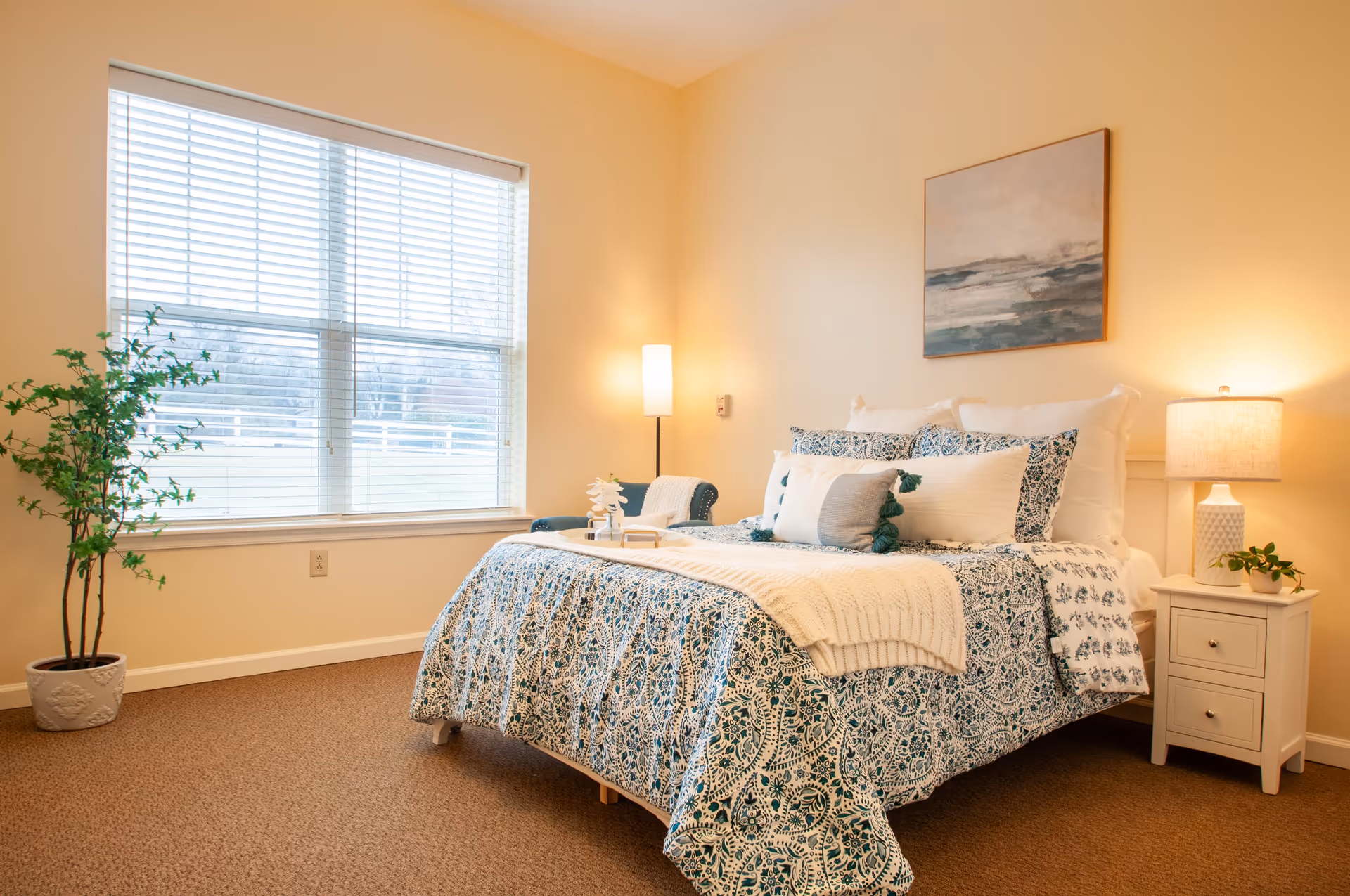 A cozy bedroom with a large window covered by white blinds, a bed with patterned blue and white bedding, multiple pillows, a white knitted throw, a white nightstand with a lamp and a small plant, a tall floor lamp, a green potted plant near the window, and a painting of a seascape on the wall.