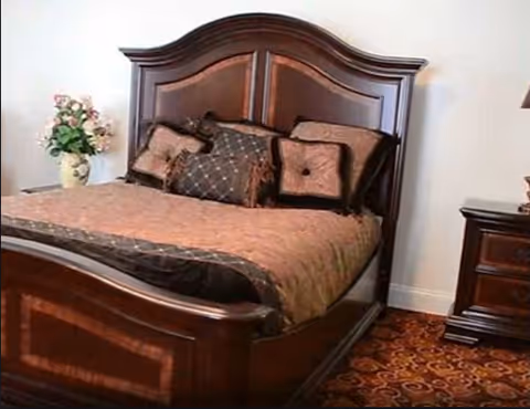 A wooden bed with an ornate headboard and footboard dressed with brown bedding and decorative pillows, next to a nightstand and a vase of flowers on patterned carpet.