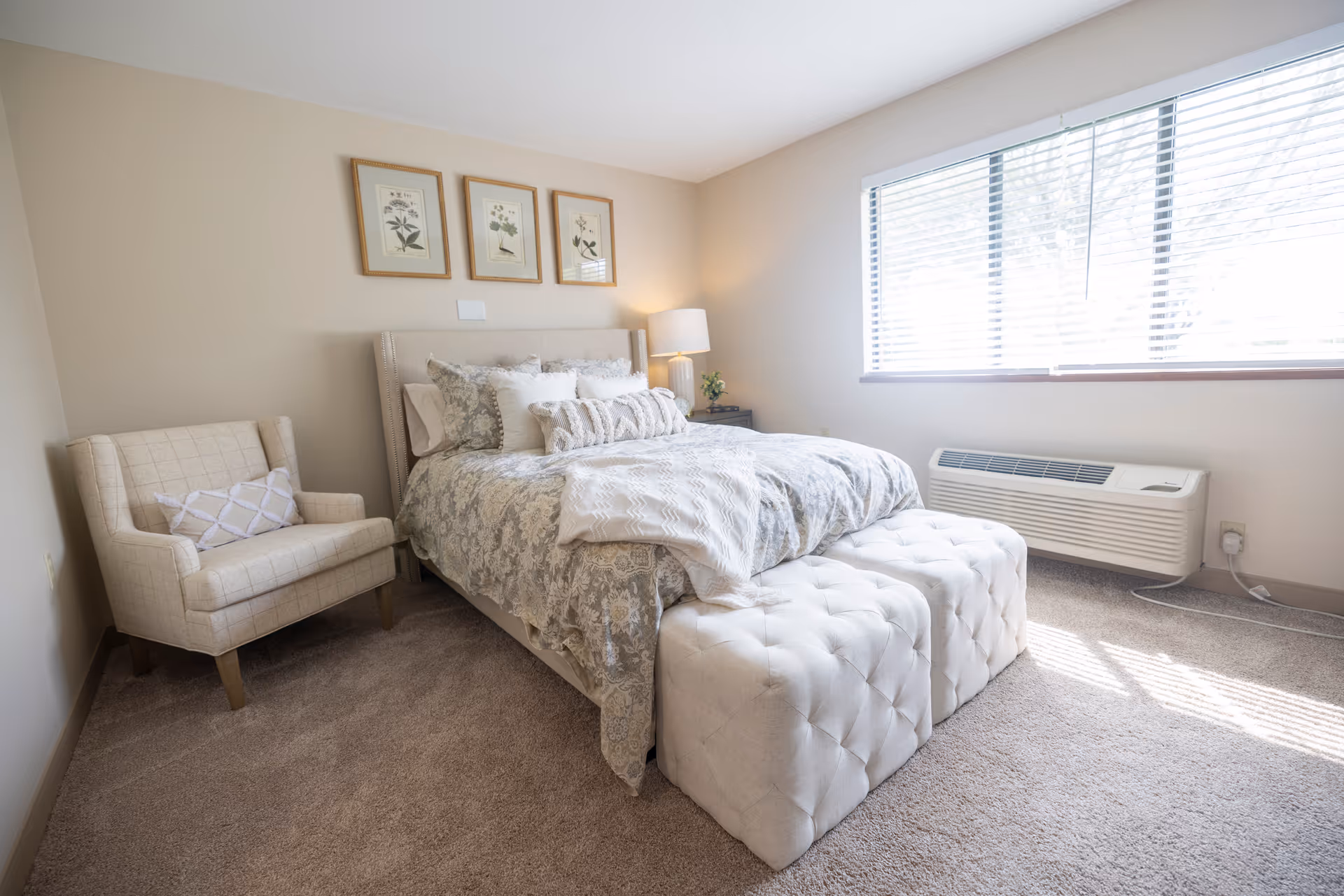 A bright bedroom with a large window letting in natural light. The room features a bed with patterned bedding and multiple pillows, a tufted bench at the foot of the bed, a beige armchair with a decorative pillow, a nightstand with a lamp and small plant, and three framed botanical prints hanging on the wall above the bed.