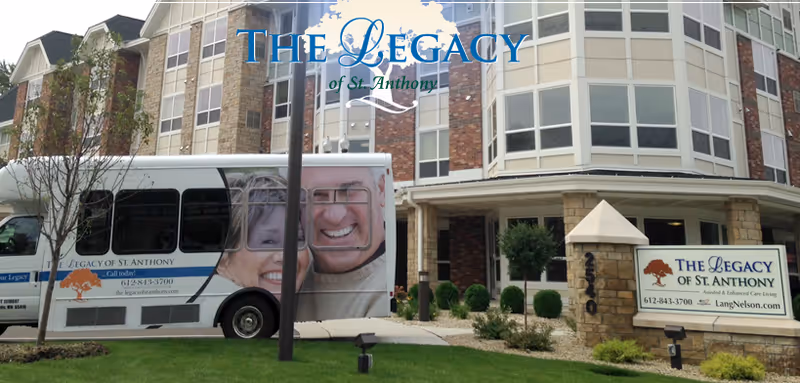 Front exterior of a senior living building with a shuttle bus parked in front and signage reading 'The Legacy of St. Anthony'.