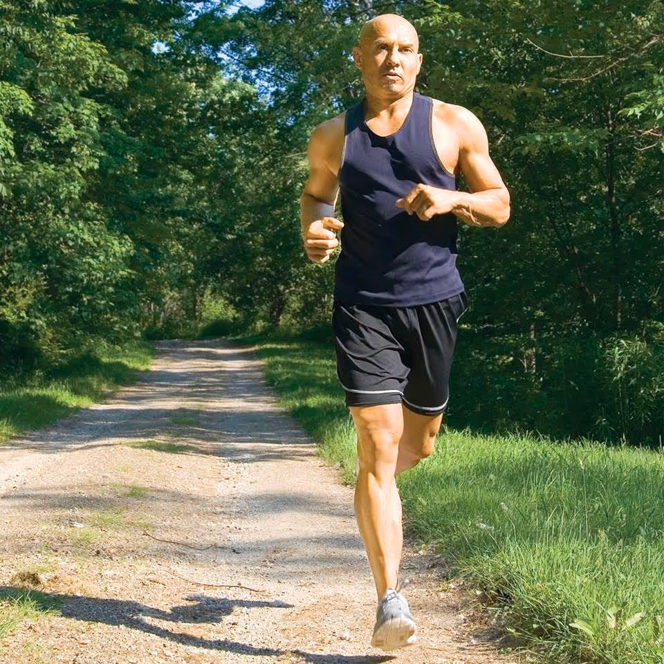 A man wearing a black tank top and black shorts jogging on a dirt path surrounded by green trees and grass on a sunny day.