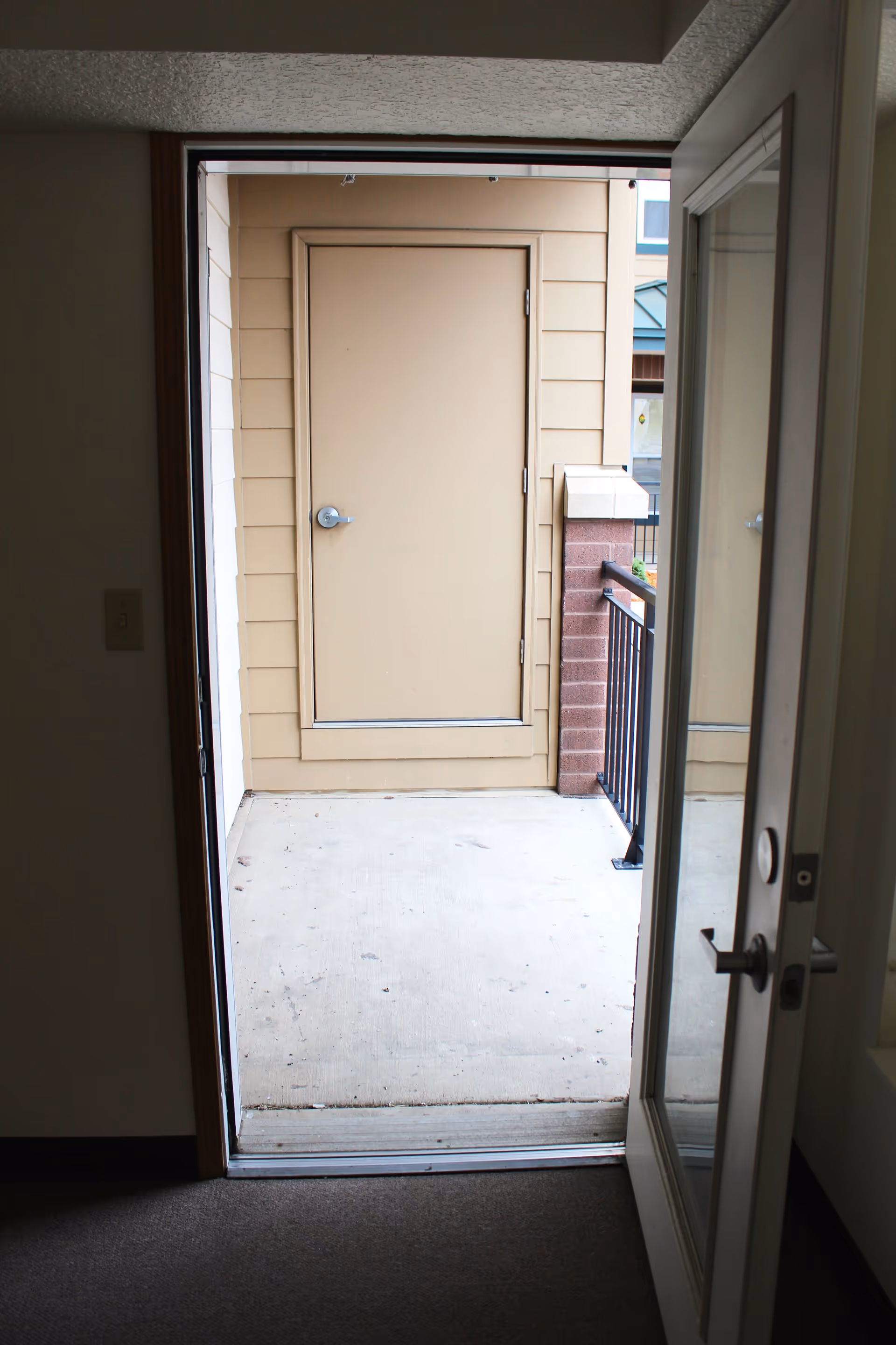 View through an open door leading to a small outdoor balcony or patio area with a closed beige door on the opposite wall, a black metal railing on the right side, and a brick pillar supporting the railing.