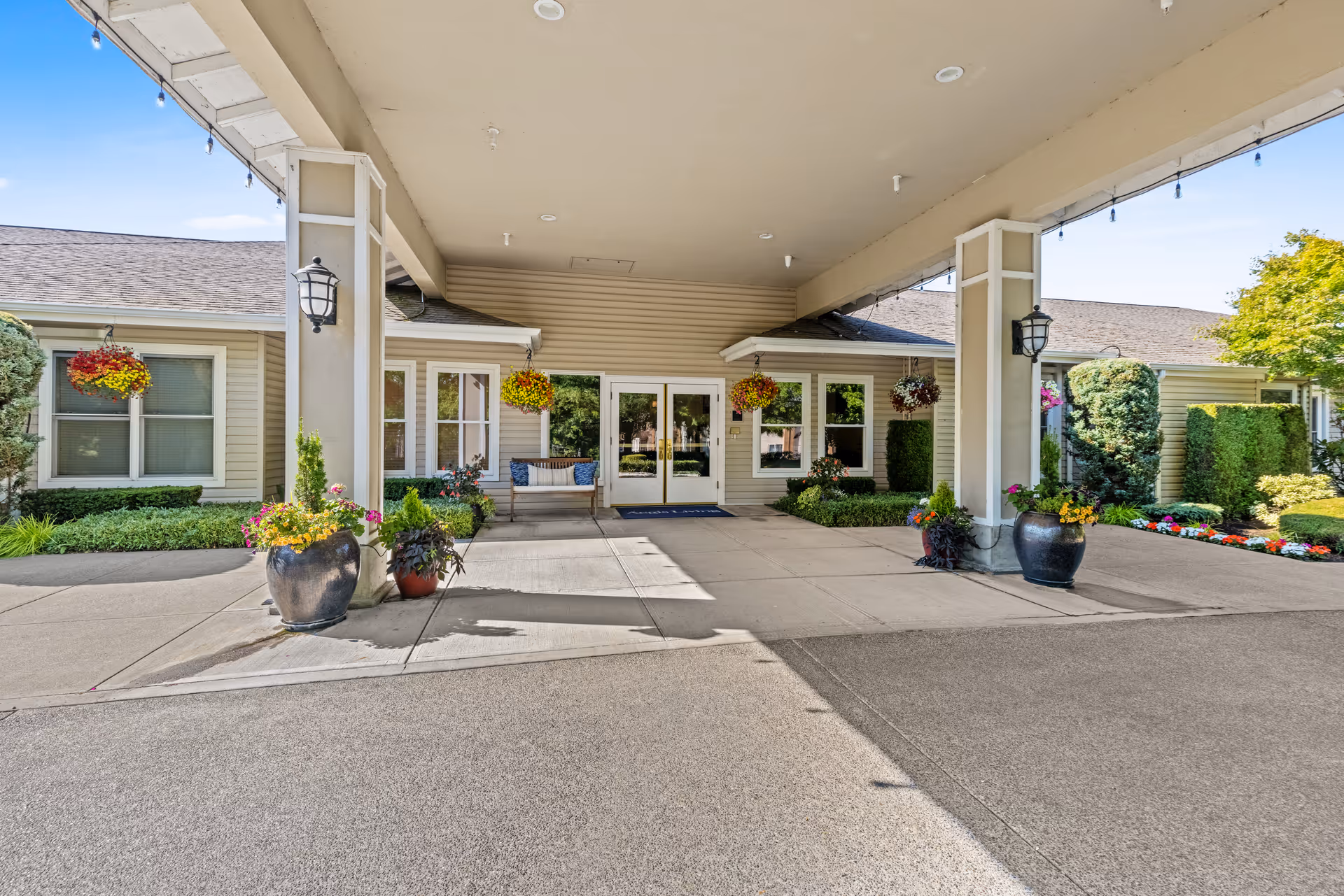 Covered entrance area of a senior living facility with hanging flower baskets, potted plants, and a bench near the double glass doors. The building exterior is beige with white trim and surrounded by well-maintained landscaping including bushes and colorful flowers.