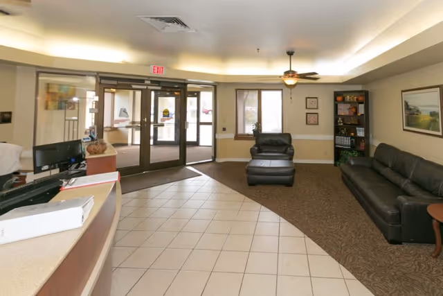 A reception lobby with a curved desk, tiled floor, seating area, and glass double entrance doors.