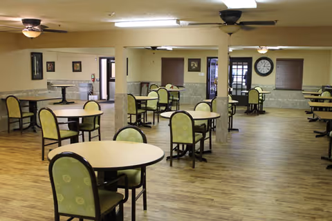 A spacious dining room with multiple round tables and green cushioned chairs arranged neatly on a wooden floor. The room has beige walls with a stone wainscot, ceiling fans, and a large clock on the far wall. There are windows with brown blinds and a door leading outside.