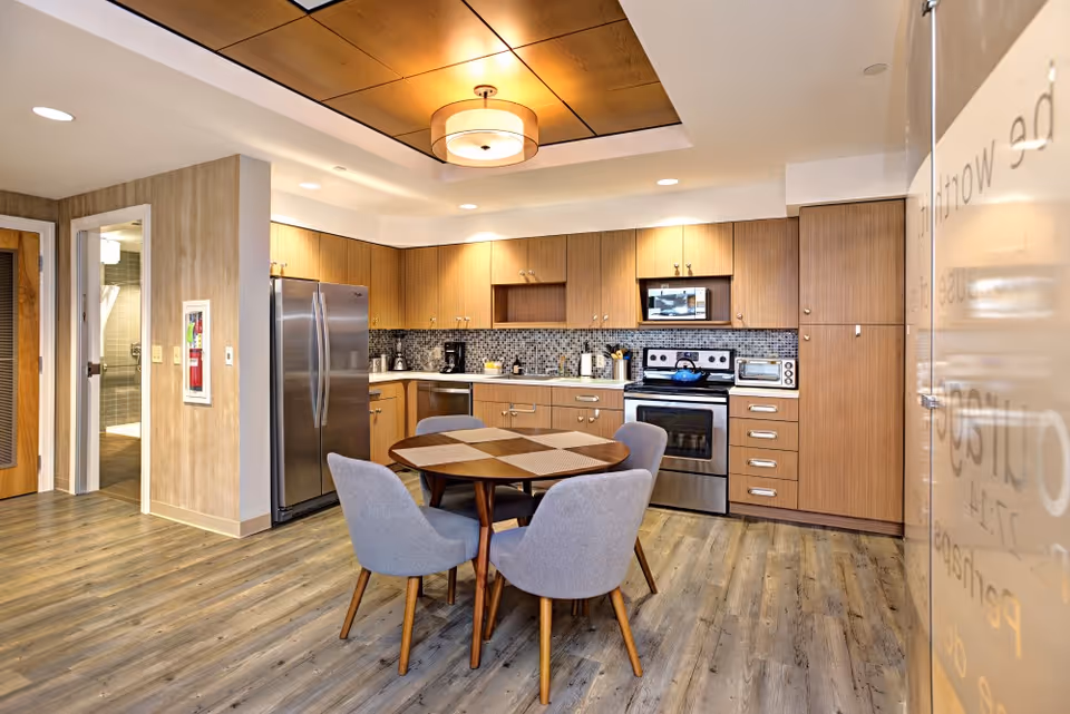 A modern kitchen area with wooden cabinets, stainless steel refrigerator, stove, microwave, and dishwasher. A round wooden table with four gray upholstered chairs is in the center. The floor has a wood-like finish, and there is a ceiling light fixture above the table. A bathroom door is visible to the left.