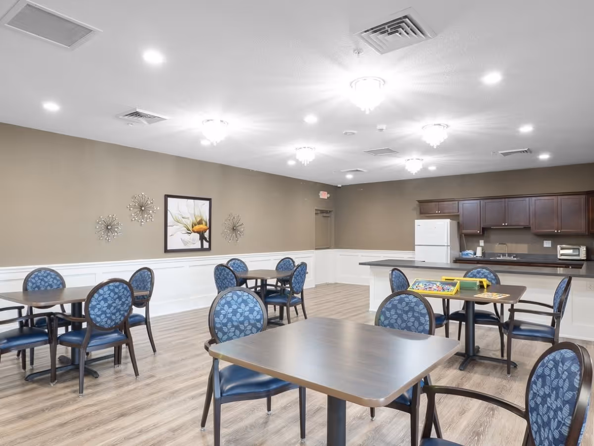 A spacious senior living dining area with round tables and blue patterned chairs, kitchenette in back.